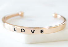 Load image into Gallery viewer, The Love Metal Cuff Bracelet in Rose Gold features a simplistic bangle with the word LOVE inscribed on the outside. Wear this bangle on its own to make a simple statement or as part of the Lainey Stack to make the perfect stack. Exclusively from Kinsley Armelle.   Details:  Style: Cuff Material: 18K Rose Gold Ion Plated Stainless Steel Size: 6.5 - 8 Inch Circumference