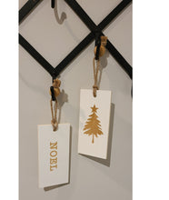 Load image into Gallery viewer, These fun and whymsical Christmas Hangers will create the perfect welcome this holiday season. With a Christmas Message on one side, and a Winter Symbol on the other, this white and gold decor will look beautiful in any room. Put it anywhere...doorknobs, walls, mantles...and you can even use it as ornaments on your tree. 2 Styles to choose from: Cheer Noel. Size: 7.5 x 3.5 x .25". Shown: Noel/Christmas Tree