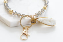 Load image into Gallery viewer, This Beaded Bracelet Keychain features a glimmering druzy white stone that will truly enchant those around you with its unique texture. The keychain has a 18k gold ion-plated stainless steel teardrop setting. It is surrounded with gold metal and grey glass beads for an elegant look. Exclusively from Kinsley Armelle. Details: Materials: 18K Yellow Gold Ion Plated Stainless Steel, druzy stone, glass and metal beads Size: 3" d Key Ring