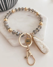 Load image into Gallery viewer, This Beaded Bracelet Keychain features a glimmering druzy white stone that will truly enchant those around you with its unique texture. The keychain has a 18k gold ion-plated stainless steel teardrop setting. It is surrounded with gold metal and grey glass beads for an elegant look. Exclusively from Kinsley Armelle. Details: Materials: 18K Yellow Gold Ion Plated Stainless Steel, druzy stone, glass and metal beads Size: 3&quot; d Key Ring
